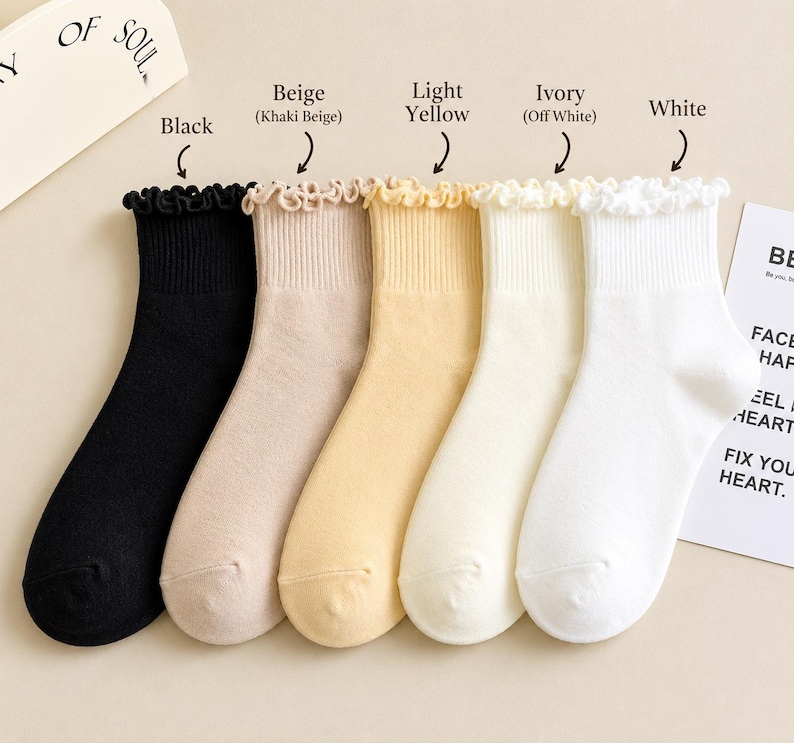 Peut inclure: Cinq paires de chaussettes &agrave; volants de couleurs vari&eacute;es : noir, beige, jaune clair, ivoire et blanc. Les chaussettes sont pr&eacute;sent&eacute;es sur un fond clair. Le texte indique "OF SOUL" et "BE FACE HAPPY FEEL HEART FIX YOUR HEART."