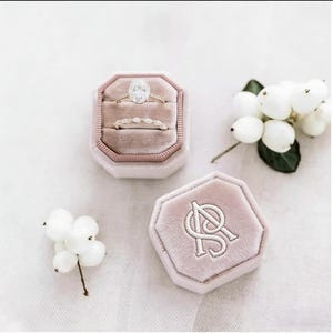 Pode incluir: Dois anéis de ouro, um com um grande diamante oval, são exibidos em uma caixa de veludo rosa. Uma segunda caixa de veludo rosa com iniciais bordadas também é mostrada. Bagas brancas e vegetação ao fundo.