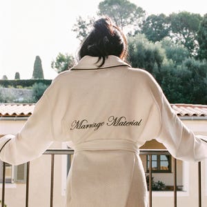 Pode incluir: Um roupão creme com acabamento preto e as palavras "Marriage Material" bordadas nas costas. O roupão é amarrado na cintura. Uma pessoa está em uma varanda com vista para as árvores.