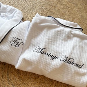 Puede incluir: Bata blanca de tejido de gofre con ribete negro y el texto bordado "Marriage Material". La bata tambi&eacute;n presenta las iniciales bordadas "FH" en negro. La bata est&aacute; doblada sobre una superficie texturizada.