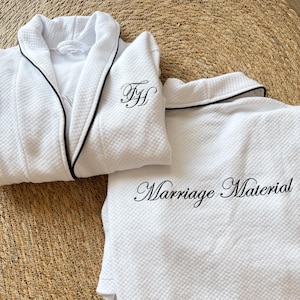 Puede incluir: Conjunto de bata y pijama acolchados blancos con ribete negro. La bata tiene iniciales bordadas y el pijama dice "Marriage Material" en escritura negra. El conjunto se muestra sobre una superficie tejida.
