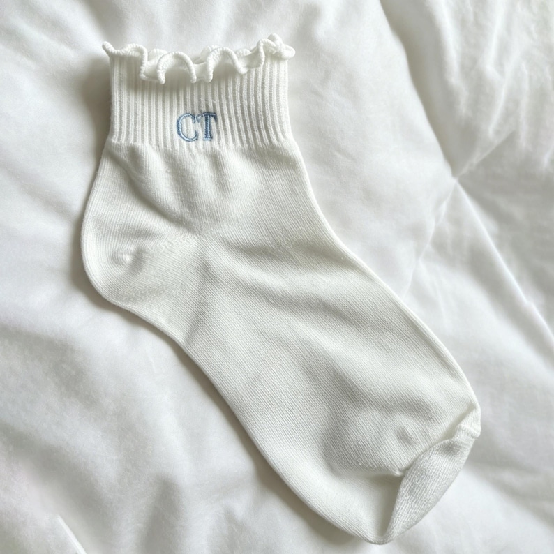 Peut inclure: Chaussette de cheville blanche avec un haut &agrave; volants et un poignet c&ocirc;tel&eacute;. La chaussette porte les lettres "CT" brod&eacute;es en bleu clair. La chaussette est faite d'une mati&egrave;re douce et textur&eacute;e.