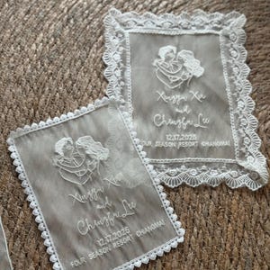 May include: Two rectangular, sheer, off-white wedding favors with white lace trim. Each favor features an embroidered illustration of a couple embracing, along with the names Xinggu Xin and Chengfre Lee, and the date 12.17.2025.