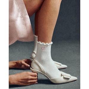 Peut inclure: Escarpins cr&egrave;me &agrave; bout pointu avec un d&eacute;tail n&oelig;ud, port&eacute;s avec des chaussettes blanches &agrave; volants brod&eacute;es d'initiales roses. Les chaussures sont tenues par une personne, avec un tissu rose clair en arri&egrave;re-plan.