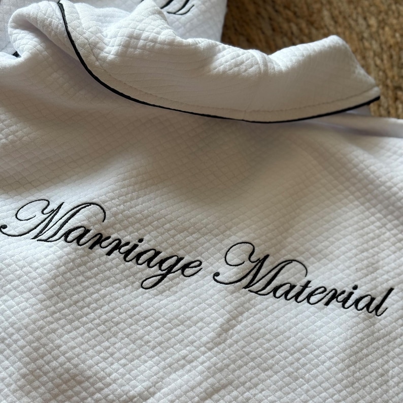 Puede incluir: Bata blanca acolchada con ribete negro y la frase bordada "Marriage Material" en elegante escritura. El cuello de la bata est&aacute; doblado y la tela tiene un patr&oacute;n texturizado en forma de diamante.