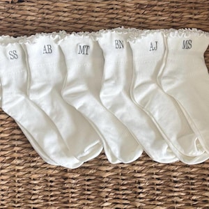 Peut inclure: Six paires de chaussettes blanches avec des volants, chacune brod&eacute;e d'initiales diff&eacute;rentes en fil gris. Les chaussettes sont dispos&eacute;es sur une surface en osier tress&eacute;. Les initiales sont SS, AB, MT, BN, AJ et MS.