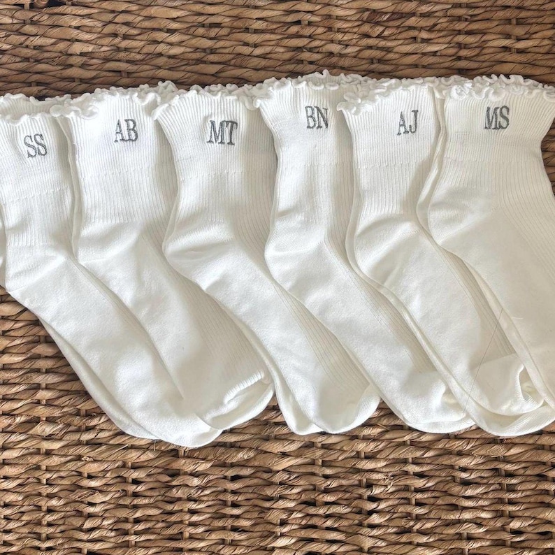 Peut inclure: Six paires de chaussettes blanches avec des volants, chacune brod&eacute;e d'initiales diff&eacute;rentes en fil gris. Les chaussettes sont dispos&eacute;es sur une surface en osier tress&eacute;. Les initiales sont SS, AB, MT, BN, AJ et MS.
