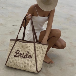 May include: A woven straw tote bag with brown leather trim and handles. The word "Bride" is embroidered in a cursive font on the front of the bag. The bag is a light beige color.