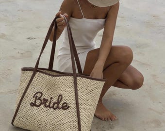 May include: A beige woven straw tote bag with brown leather trim and handles. The word "Bride" is embroidered in a cursive font on the front. The bag is a light beige colour.