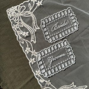 May include: A delicate, white lace wedding handkerchief with embroidered floral and bow details. The handkerchief features two rectangular embroidered labels that read "Bride" and "Groom" in cursive script.