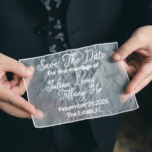 Może przedstawiać: Przezroczyste ogłoszenie ślubne z białym haftowanym tekstem. Tekst brzmi "Save The Date", a następnie imiona "Julian Leong" i "Tiffany Ho", data "29 listopada 2025" oraz lokalizacja "The Estate, KL."