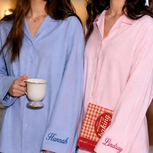 Op de afbeelding: Een lichtblauw pyjamashirt met knopen en de naam Hannah in donkerblauw geborduurd op de manchet, en een lichtroze pyjamashirt met knopen en de naam Lindsay in donkerroze geborduurd op de manchet, beide voorzien van een reverskraag.