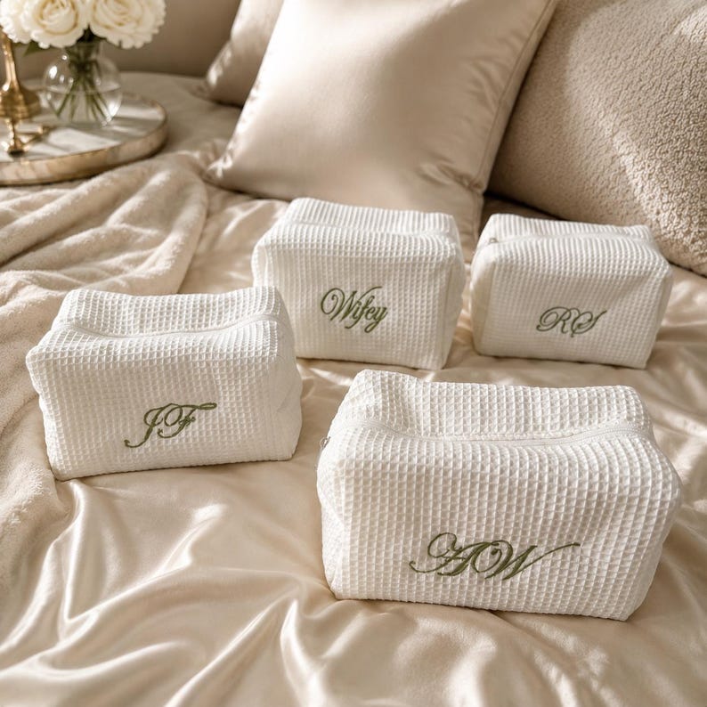 May include: Four white waffle-textured fabric pouches with zippered closures. Each pouch features custom green embroidered lettering, including the text Wifey, RS, JF, and AW, arranged on a soft, draped fabric surface with a subtle sheen.