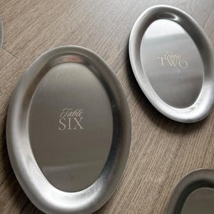 Może przedstawiać: Kilka owalnych tac do serwowania ze szczotkowanego metalu. Tace mają srebrne wykończenie i są wygrawerowane słowami "Table SIX" i "Table TWO". Tace leżą na drewnianej powierzchni.