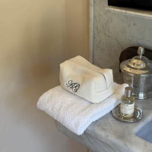 Puede incluir: Un neceser blanco con textura de gofre con las iniciales "AB" bordadas descansa sobre una toalla blanca doblada. Un pequeño frasco de perfume se encuentra sobre una bandeja plateada, junto a un recipiente plateado con tapa.