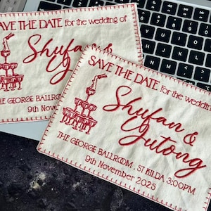 Op de afbeelding: Drie crèmekleurige stoffen save-the-date kaarten met rood geborduurde tekst. De kaarten luiden "SAVE THE DATE for the wedding of Shufan & Jutong" met datum en locatie. Een champagne toren grafiek is ook geborduurd.
