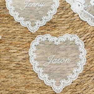Può includere: Primo piano di segnaposto in pizzo a forma di cuore con i nomi "Jenny" e "Jason" ricamati in corsivo. Il delicato pizzo bianco è posto su uno sfondo tessuto dai toni naturali, ideale per matrimoni o eventi speciali.