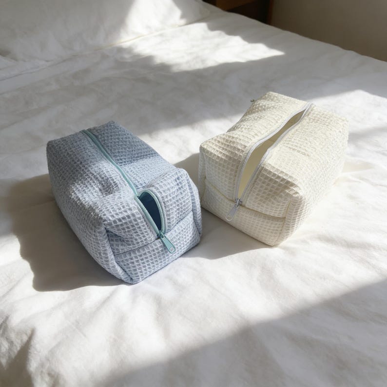 May include: Two rectangular toiletry bags with a waffle-textured fabric. One bag is light blue with a matching zipper, and the other is off-white with a white zipper. Both bags feature a top-opening design with a single zipper pull closure.
