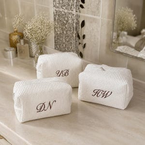 May include: Three white rectangular toiletry bags made of a textured waffle-weave fabric. Each bag features a silver zipper closure and is personalized with brown embroidered initials in a cursive script, specifically reading YB, DN, and HW.