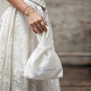 Könnte beinhalten: Ein weißer Satinbeutel mit Knotenhenkel, der das Wort bridesmaid in hellblauer Schreibschrift gestickt zeigt. Die Tasche hat eine glatte, glänzende Oberfläche und einen gerafften Verschluss, gehalten von einer Hand mit Perlenring und dünnem Armband.