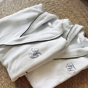 Puede incluir: Dos albornoces blancos de tejido de gofre con ribete negro e iniciales bordadas. Las batas est&aacute;n dobladas, mostrando la tela texturizada y el dise&ntilde;o elegante. Las iniciales est&aacute;n en una fuente elegante.