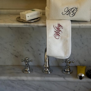 May include: A bathroom setting with a white waffle-weave robe, embroidered with black initials, and a white towel embroidered with the word "Wifey" in burgundy. A bar of soap rests on a silver dish, with a tube of yellow cream and a brush also present.
