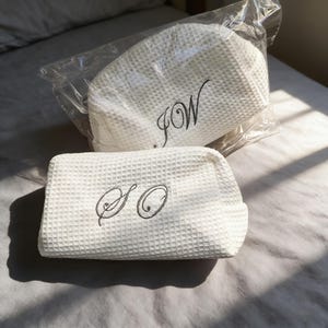 May include: Two white waffle-textured fabric pouches with dark grey embroidered lettering. One pouch is inside a clear plastic bag and features the letters JW, while the other is outside the bag and features the letters SO in a cursive script font.