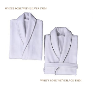 Puede incluir: Dos albornoces blancos con cuello chal. Uno tiene ribete plateado, el otro negro. El texto "WHITE ROBE WITH SILVER TRIM" y "WHITE ROBE WITH BLACK TRIM" se muestra encima y debajo de los albornoces.