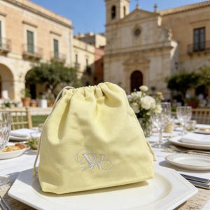 Può includere: Un sacchetto con coulisse giallo pallido con iniziali ricamate bianche, appoggiato su un piatto bianco. La borsa ha un aspetto morbido e strutturato e un cordoncino di colore naturale. Lo sfondo mostra un'ambientazione formale per la ristorazione all'aperto con un edificio storico.