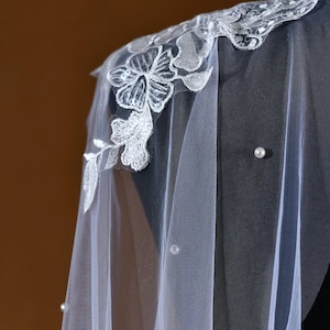 Pu&ograve; includere: Un velo bianco trasparente con delicati ricami floreali lungo il bordo. Il velo presenta intricati dettagli in pizzo bianco ed &egrave; adornato da perle sparse. Il velo &egrave; drappeggiato su una superficie scura, mostrando il suo design elegante.