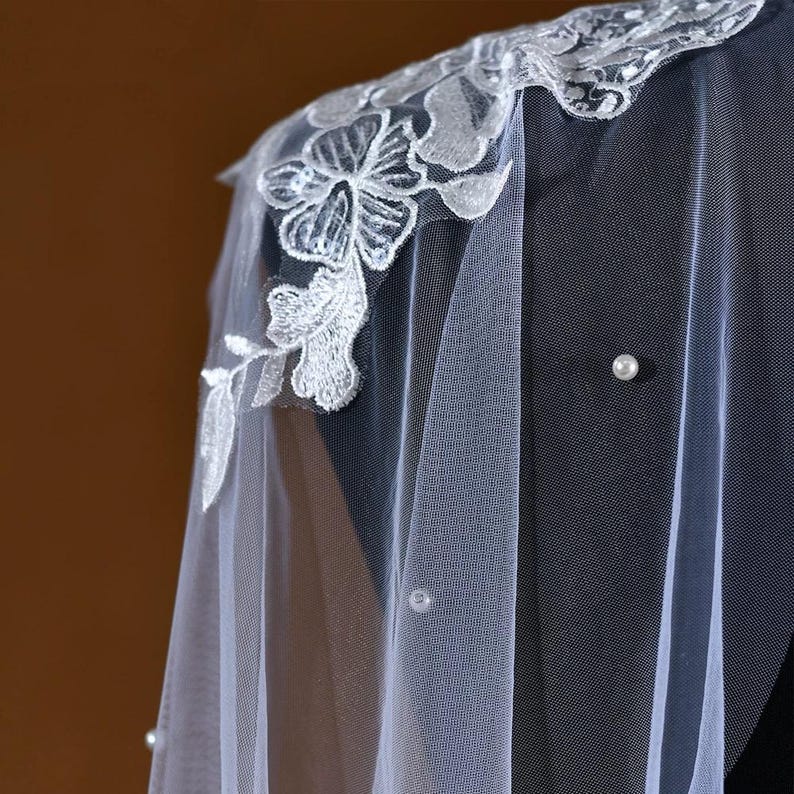 Pu&ograve; includere: Un velo bianco trasparente con delicati ricami floreali lungo il bordo. Il velo presenta intricati dettagli in pizzo bianco ed &egrave; adornato da perle sparse. Il velo &egrave; drappeggiato su una superficie scura, mostrando il suo design elegante.