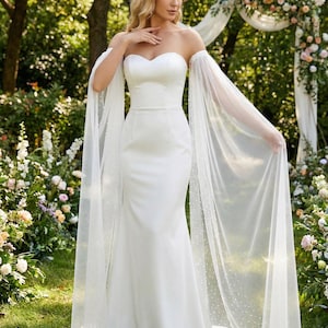 May include: A white strapless wedding dress with a fitted bodice and a flowing mermaid skirt. The dress features sheer, dotted sleeves that drape elegantly. The bride is standing in a garden setting with floral decorations.
