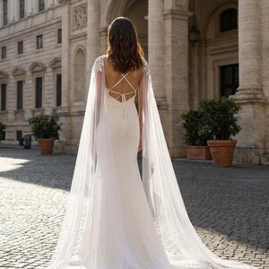 Pu&ograve; includere: Abito da sposa bianco con un lungo strascico e una mantella trasparente ornata di piccole perle. L'abito presenta una schiena scoperta con spalline incrociate. La sposa &egrave; in piedi di fronte a un edificio classico con archi e colonne.