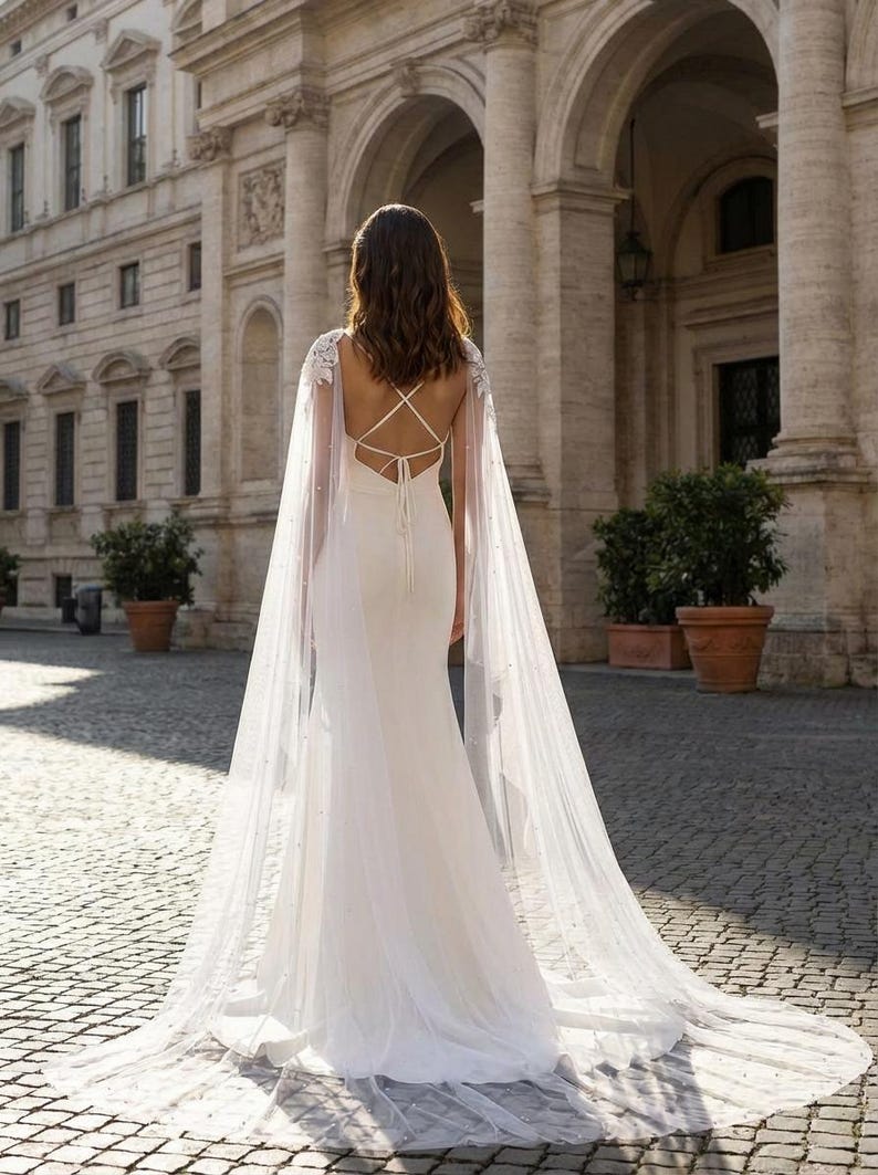 Pu&ograve; includere: Abito da sposa bianco con un lungo strascico e una mantella trasparente ornata di piccole perle. L'abito presenta una schiena scoperta con spalline incrociate. La sposa &egrave; in piedi di fronte a un edificio classico con archi e colonne.
