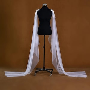 Pu&ograve; includere: Una mantella da sposa bianca con delicate decorazioni floreali sulle spalle. La mantella &egrave; realizzata in un tessuto trasparente e fluido che scende fino a terra. La mantella &egrave; esposta su un manichino nero su sfondo marrone.
