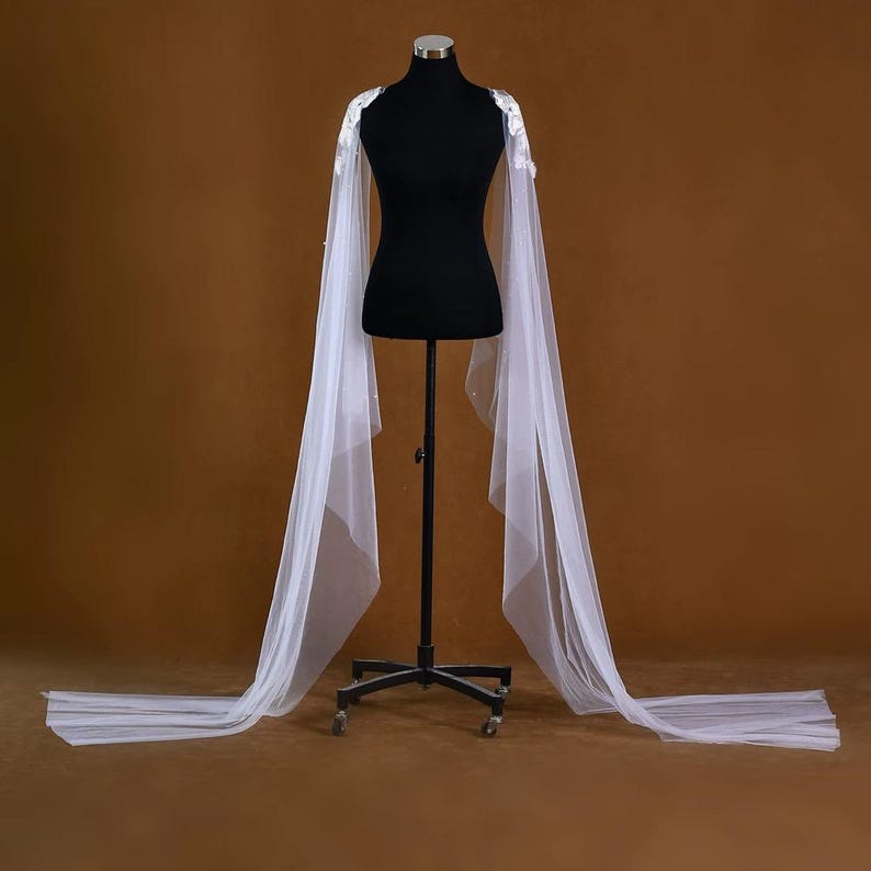 Pu&ograve; includere: Una mantella da sposa bianca con delicate decorazioni floreali sulle spalle. La mantella &egrave; realizzata in un tessuto trasparente e fluido che scende fino a terra. La mantella &egrave; esposta su un manichino nero su sfondo marrone.