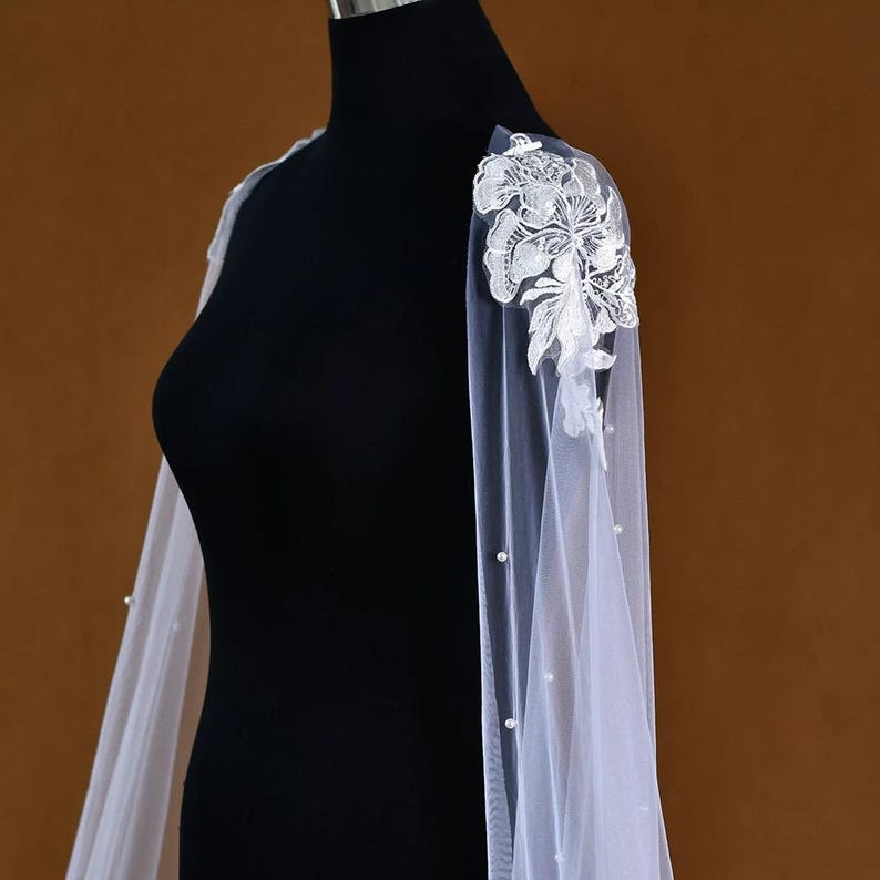Pu&ograve; includere: Una mantella da sposa bianca trasparente con delicati dettagli floreali in pizzo sulle spalle. La mantella presenta un design fluido con piccoli accenti di perle lungo i bordi. La mantella &egrave; esposta su un manichino nero su sfondo neutro.
