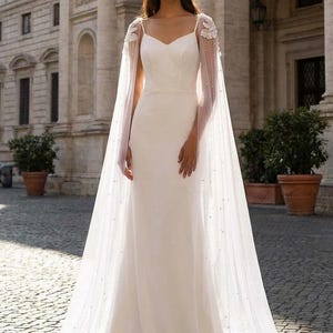 Pu&ograve; includere: Un abito da sposa bianco con scollatura a cuore e gonna fluida. L'abito &egrave; abbinato a un mantello trasparente ornato da delicati ricami floreali e dettagli perlati sparsi. Il mantello drappeggia con grazia, aggiungendo un tocco etereo all'ensemble nuziale.
