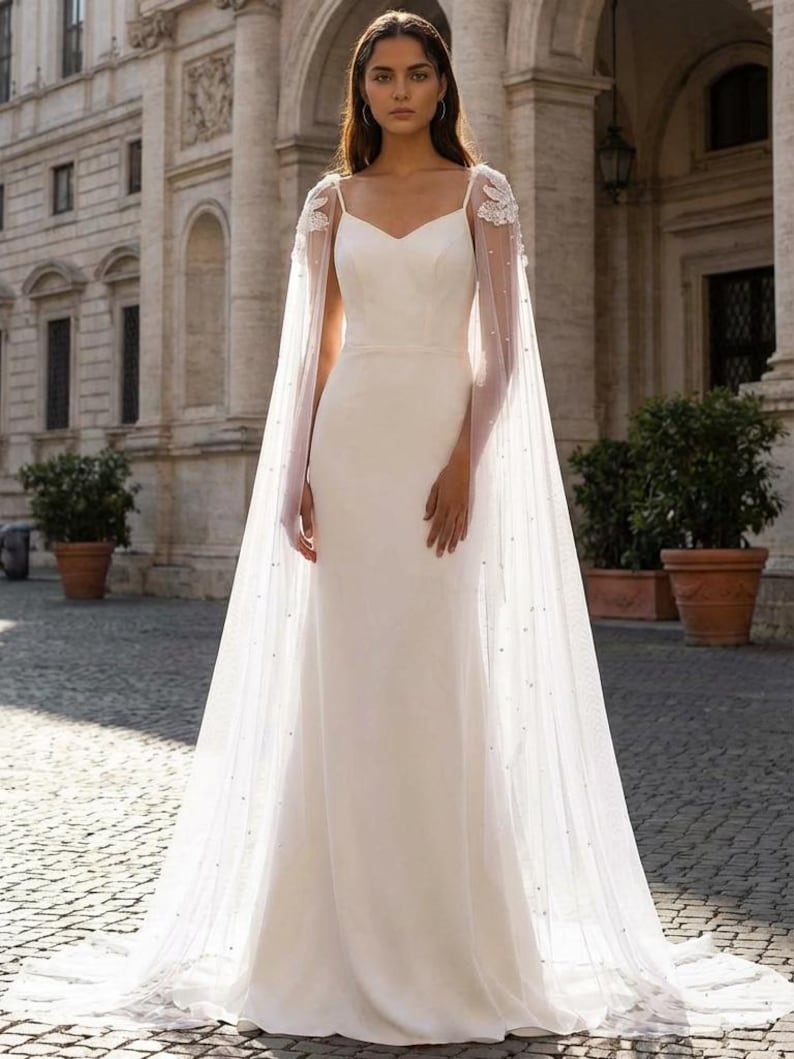 Pu&ograve; includere: Un abito da sposa bianco con scollatura a cuore e gonna fluida. L'abito &egrave; abbinato a un mantello trasparente ornato da delicati ricami floreali e dettagli perlati sparsi. Il mantello drappeggia con grazia, aggiungendo un tocco etereo all'ensemble nuziale.