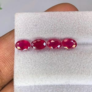 Può includere: Quattro rubini rosso intenso dal taglio ovale sono disposti in una fila orizzontale. Ogni gemma presenta una superficie sfaccettata che riflette la luce, mostrando una vibrante tonalità cremisi. Le pietre sono posizionate su un cuscinetto in tessuto grigio chiaro.