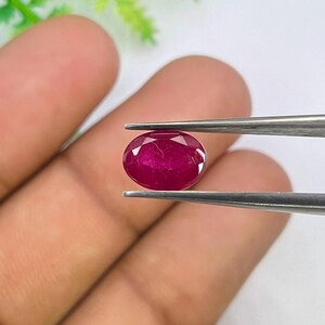 Könnte beinhalten: Ein tiefroter, oval geschliffener Edelstein mit facettierter Oberfläche, der von einer Metallpinzette gehalten wird. Der Stein zeigt einen satten, kräftigen Karmesinton und eine polierte Oberfläche, die das Licht auf den Facetten reflektiert.