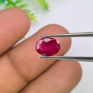 Puede incluir: Una gema de rubí de corte ovalado con una superficie facetada que refleja la luz. La piedra de color rojo intenso está sujeta por unas pinzas de metal plateado, mostrando su calidad translúcida y su corte geométrico preciso sobre un fondo.
