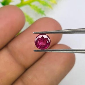 Può includere: Una pietra preziosa rotonda, sfaccettata e di colore rosa intenso, tenuta da una pinzetta metallica. La pietra presenta un taglio brillante con molteplici sfaccettature triangolari che riflettono la luce, creando un profilo cromatico vibrante.