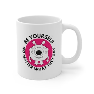 Könnte beinhalten: Weiße Keramiktasse mit einem Cartoon-Monster-Design in einem rosa Kreis. Der Text "BE YOURSELF NO MATTER WHAT THEY SAY" umgibt das Monster. Die Tasse hat einen gebogenen Griff.