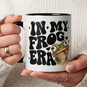 May include: A white ceramic mug with a black handle and rim, featuring the black text "IN MY FROG ERA" in a bold, wavy font, accompanied by small star and heart icons and a detailed illustration of a green frog with large eyes.