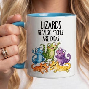 May include: A white ceramic mug with a light blue handle and interior. The mug features the black text "LIZARDS BECAUSE PEOPLE ARE DICKS" above a colourful illustration of five cartoon lizards in blue, green, purple, orange, and yellow.
