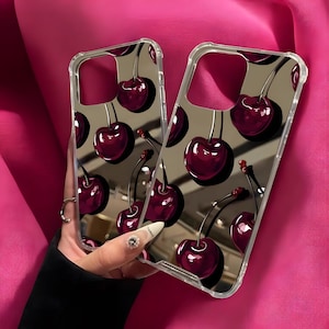 Op de afbeelding: Transparante telefoonhoesjes met een herhaald patroon van glanzende, donkerrode kersen. De kersen hebben groene steeltjes en zijn geplaatst tegen een reflecterende, grijze achtergrond. De hoesjes worden tegen een roze achtergrond gehouden.