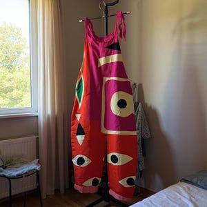 May include: A striking jumpsuit in bold colours. The garment displays a mix of red, pink, and black geometric shapes, and eye-shaped patterns on the legs. It hangs on a black clothes rack in a room with a window, showcasing a unique design.