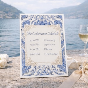 Könnte beinhalten: Eine dekorative Hochzeitsplan-Karte mit blauem und weißem floralem Design. Die Karte zeigt den Text "The Celebration Schedule" und die Veranstaltungszeiten. Ein Glas Weißwein und Blumen sind im Hintergrund.