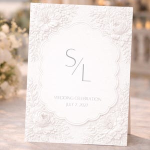 May include: A white wedding invitation with embossed floral details and a scalloped border. The invitation features the initials "S/L" and the text "WEDDING CELEBRATION JULY 7, 2027". The card is standing upright.